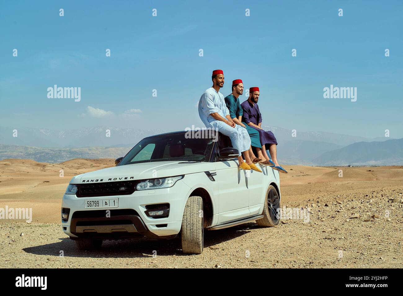 Trois hommes en tenue traditionnelle assis au sommet d'un SUV blanc sur fond d'un ciel bleu clair et d'un paysage aride, Maroc Banque D'Images