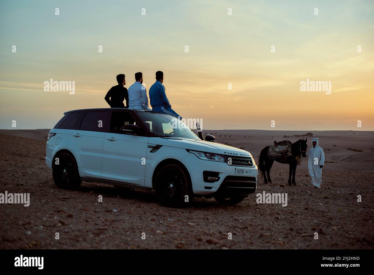 Deux hommes sont assis au sommet d'un SUV blanc dans un désert pendant le coucher du soleil, avec un troisième homme debout à côté d'un cheval en arrière-plan. Banque D'Images