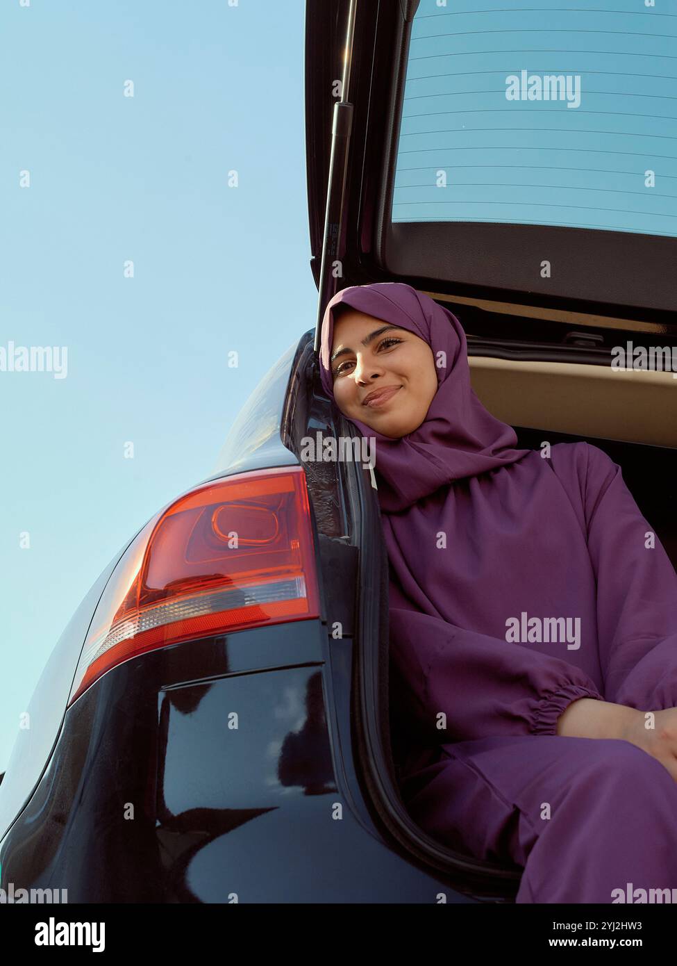 Femme souriante en tenue violette assise dans le coffre d'une voiture avec un ciel bleu clair en arrière-plan, Maroc Banque D'Images