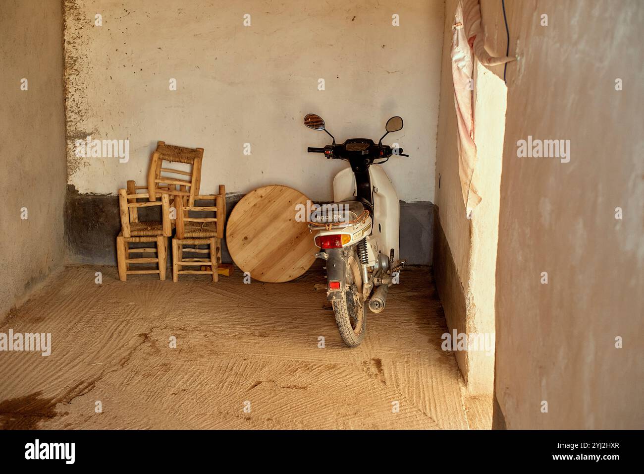 Un scooter blanc garé à l'intérieur d'une pièce rustique avec un sol recouvert de paille, à côté d'une chaise en bois et d'une table circulaire appuyée contre le mur, Maroc Banque D'Images