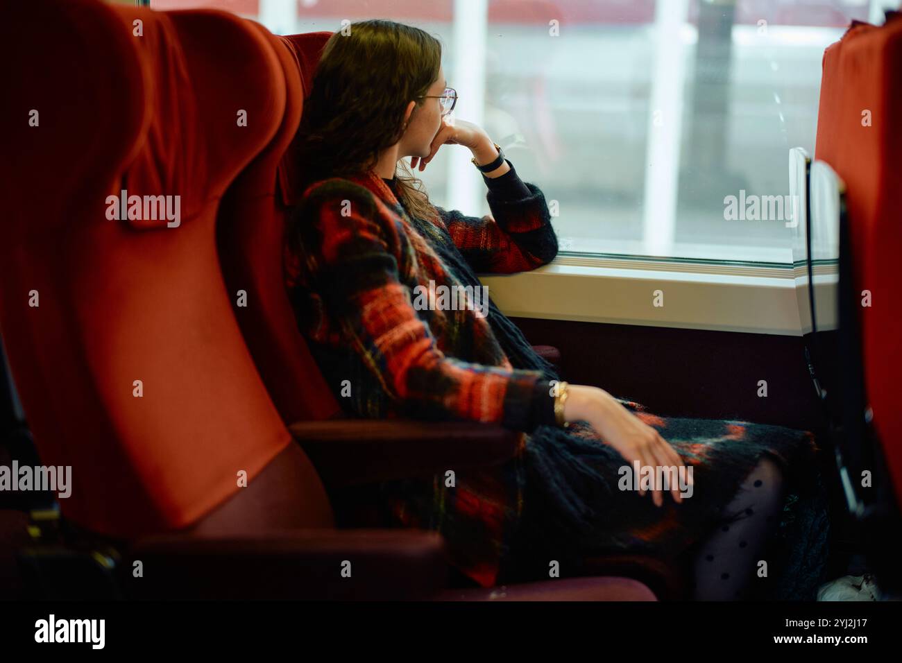 Femme en lunettes assise confortablement dans un siège de train rouge, regardant par la fenêtre profondément dans la pensée. Banque D'Images