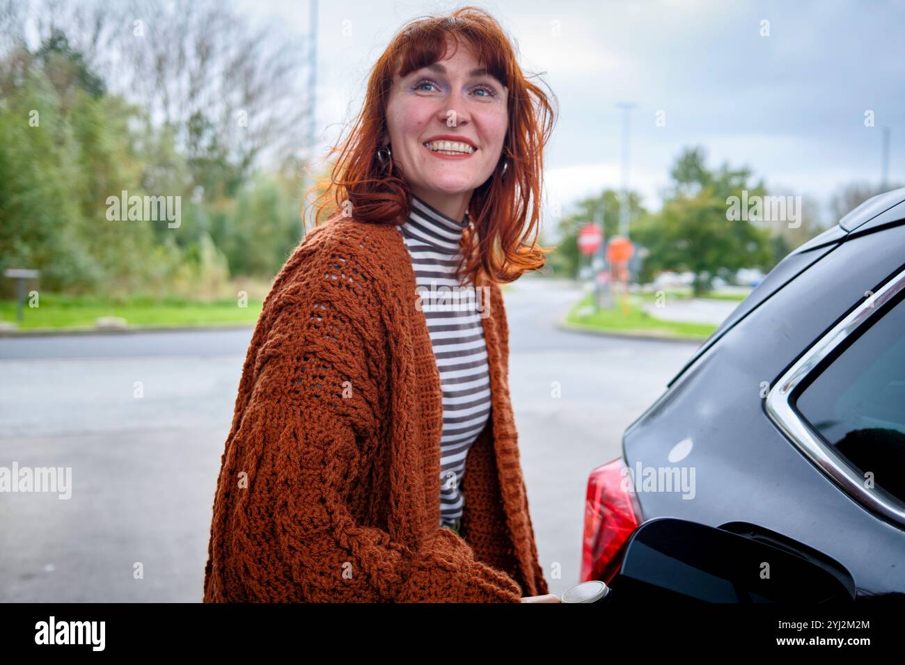 Femme souriante portant un cardigan marron et une chemise rayée debout à côté d'une voiture par temps couvert, Bruxelles, Belgique Banque D'Images