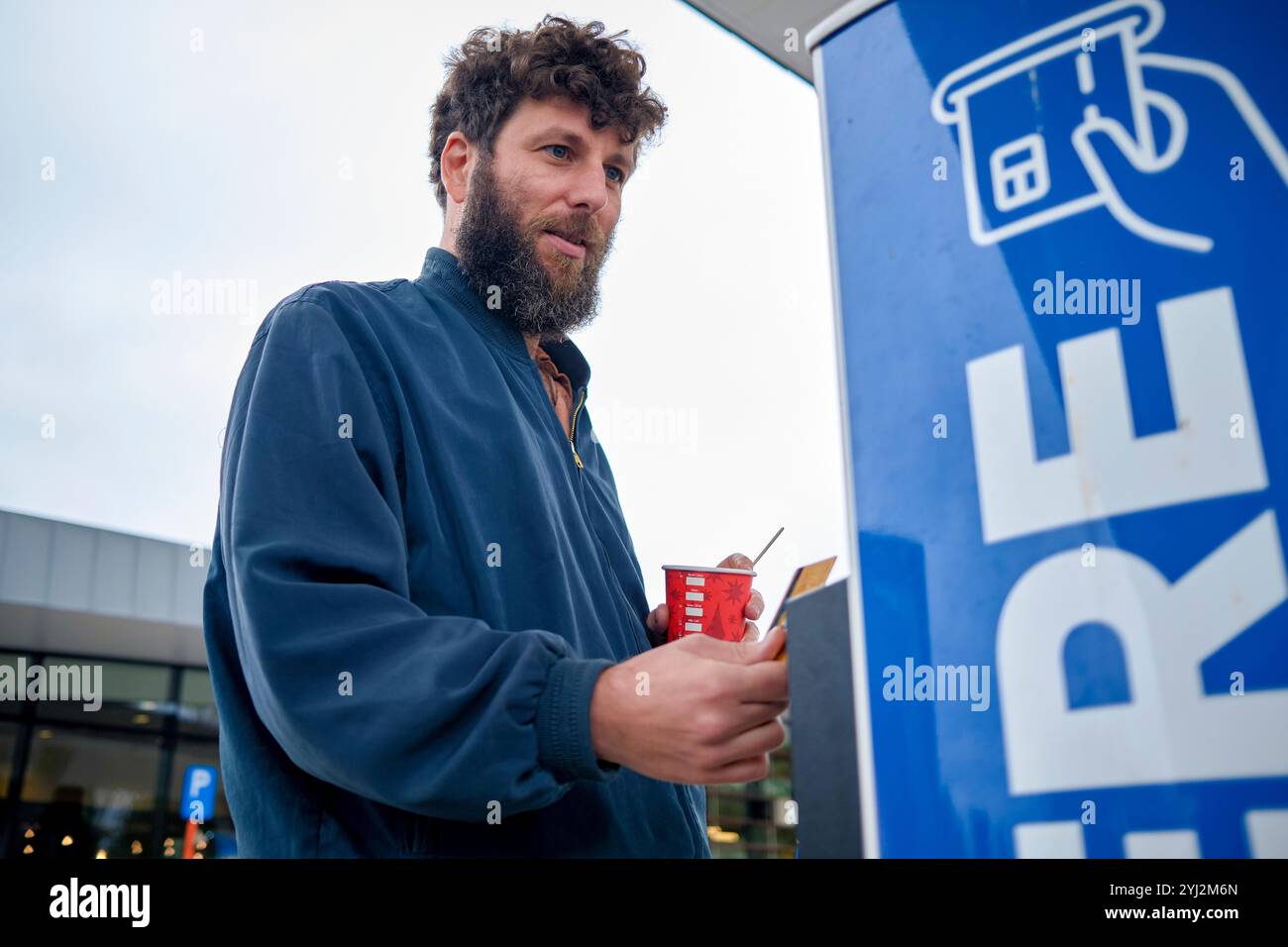 Un homme se tient à l'extérieur près d'une borne de recharge de véhicule électrique, tenant une tasse à café et un smartphone, avec un bâtiment moderne en arrière-plan, Bruxelles, Belgique Banque D'Images