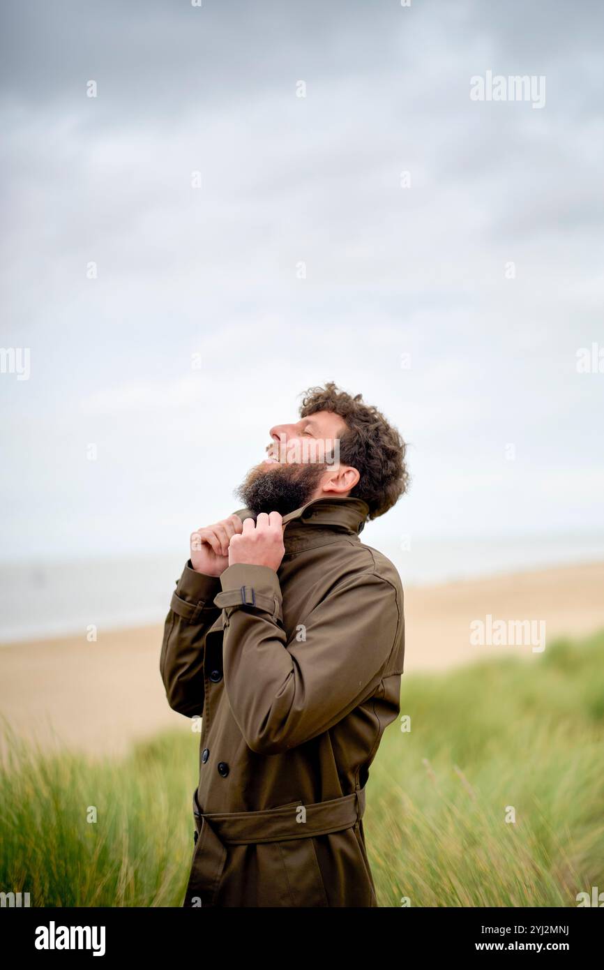 Homme dans un trench-coat brun se tient dans un champ herbeux contre un ciel nuageux, ajustant son col et regardant vers le haut, Belgique Banque D'Images