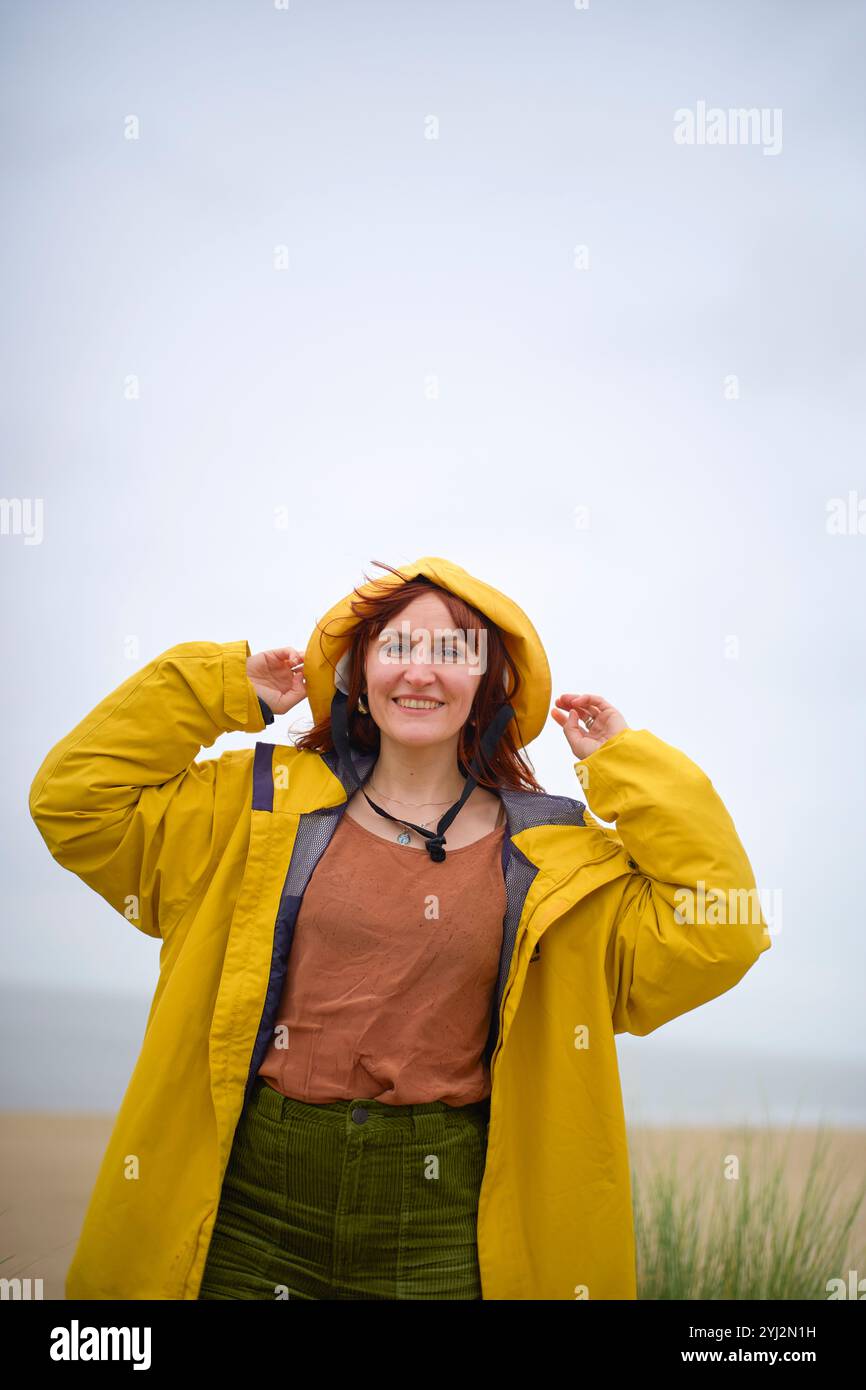 Femme souriante dans une veste jaune et un chapeau debout sur une plage par un jour nuageux, Belgique Banque D'Images