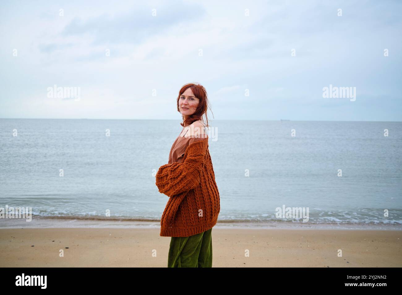 Femme debout sur la plage tenant un grand sac tricoté, regardant par-dessus l'épaule avec l'océan en arrière-plan, Belgique Banque D'Images