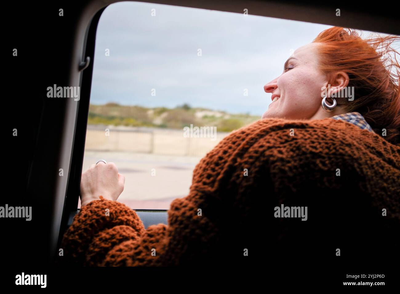 Une femme joyeuse aux cheveux roux profitant de la brise à travers une fenêtre de voiture, les yeux fermés et un large sourire sur son visage, Belgique Banque D'Images