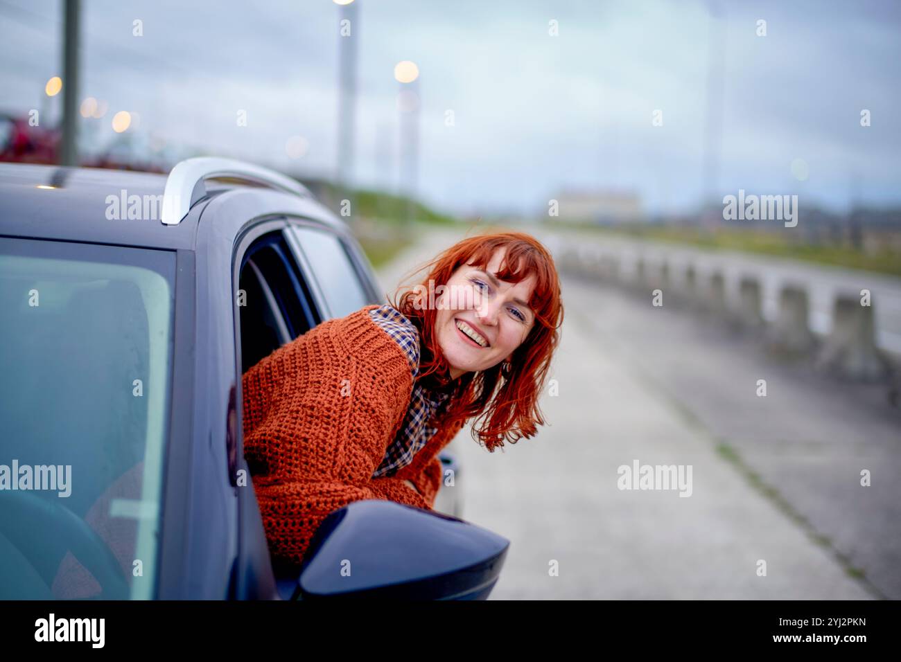 Femme souriante avec les cheveux roux jetant un coup d'œil par une fenêtre de voiture par un jour nuageux. Banque D'Images