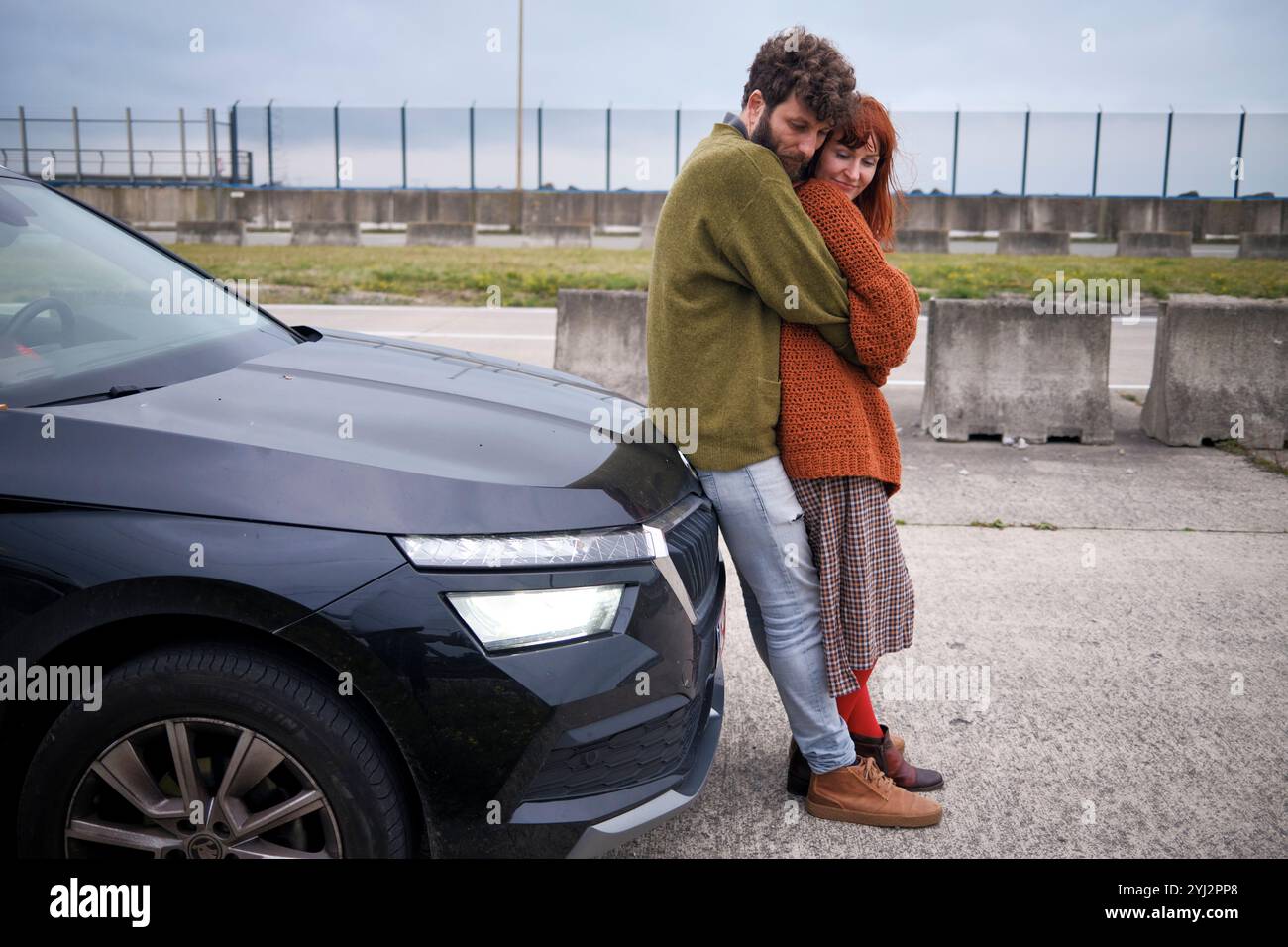 Couple embrassant à côté d'une voiture noire par une journée nuageuse avec un fond industriel, la Belgique Banque D'Images