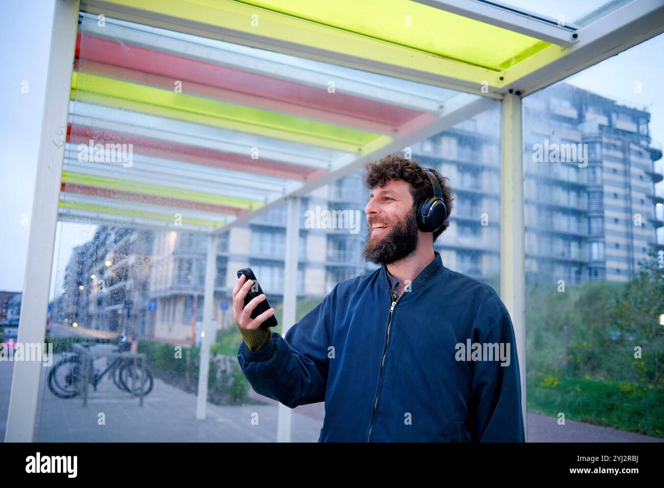 Homme souriant avec des écouteurs tenant un smartphone à un arrêt de bus coloré pendant le crépuscule, Belgique Banque D'Images