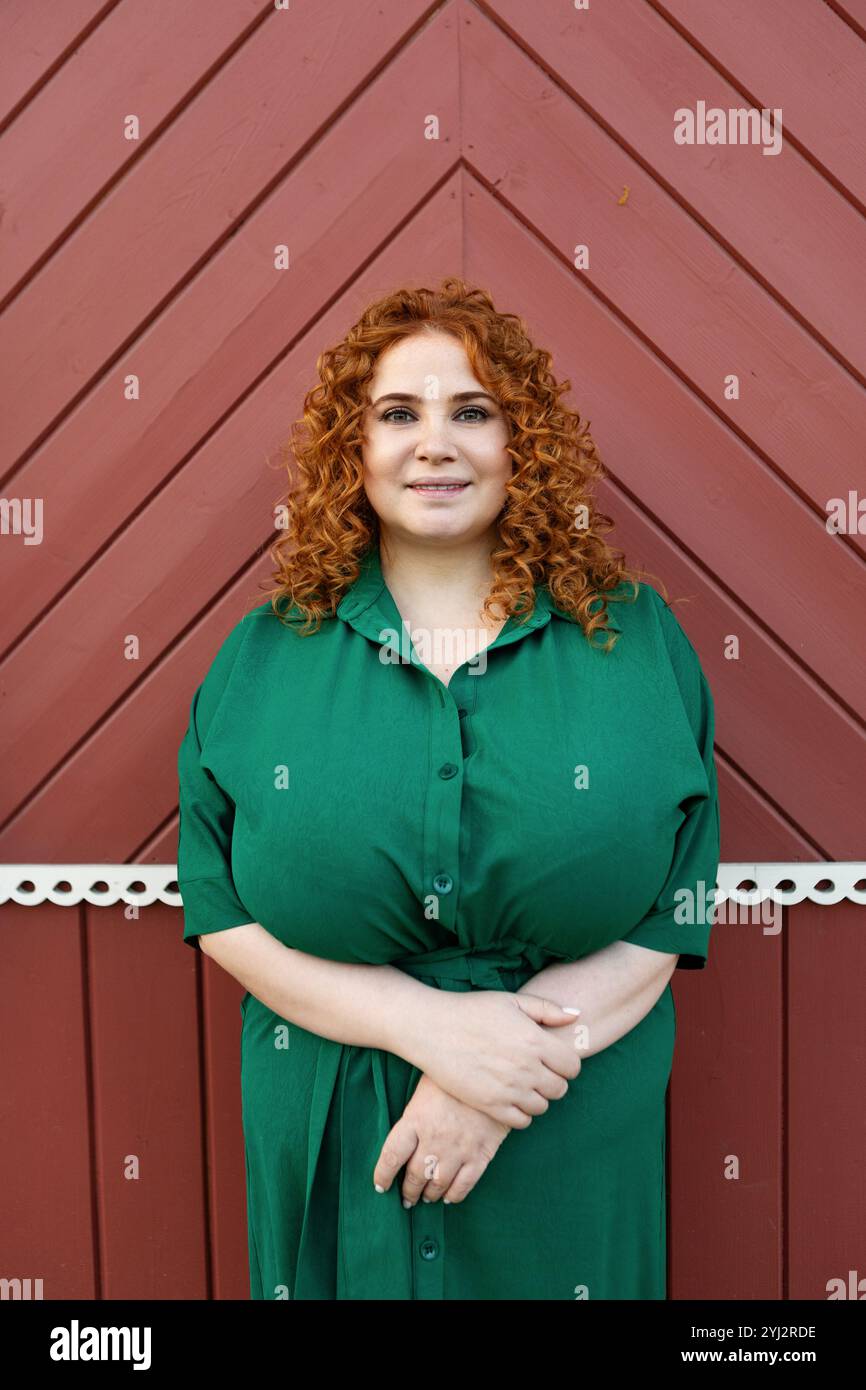 Portrait plus taille belle femme avec cheveux rouges bouclés dans la robe verte regardant la caméra contre fond rouge coloré de mur, beauté et confiance ou Banque D'Images
