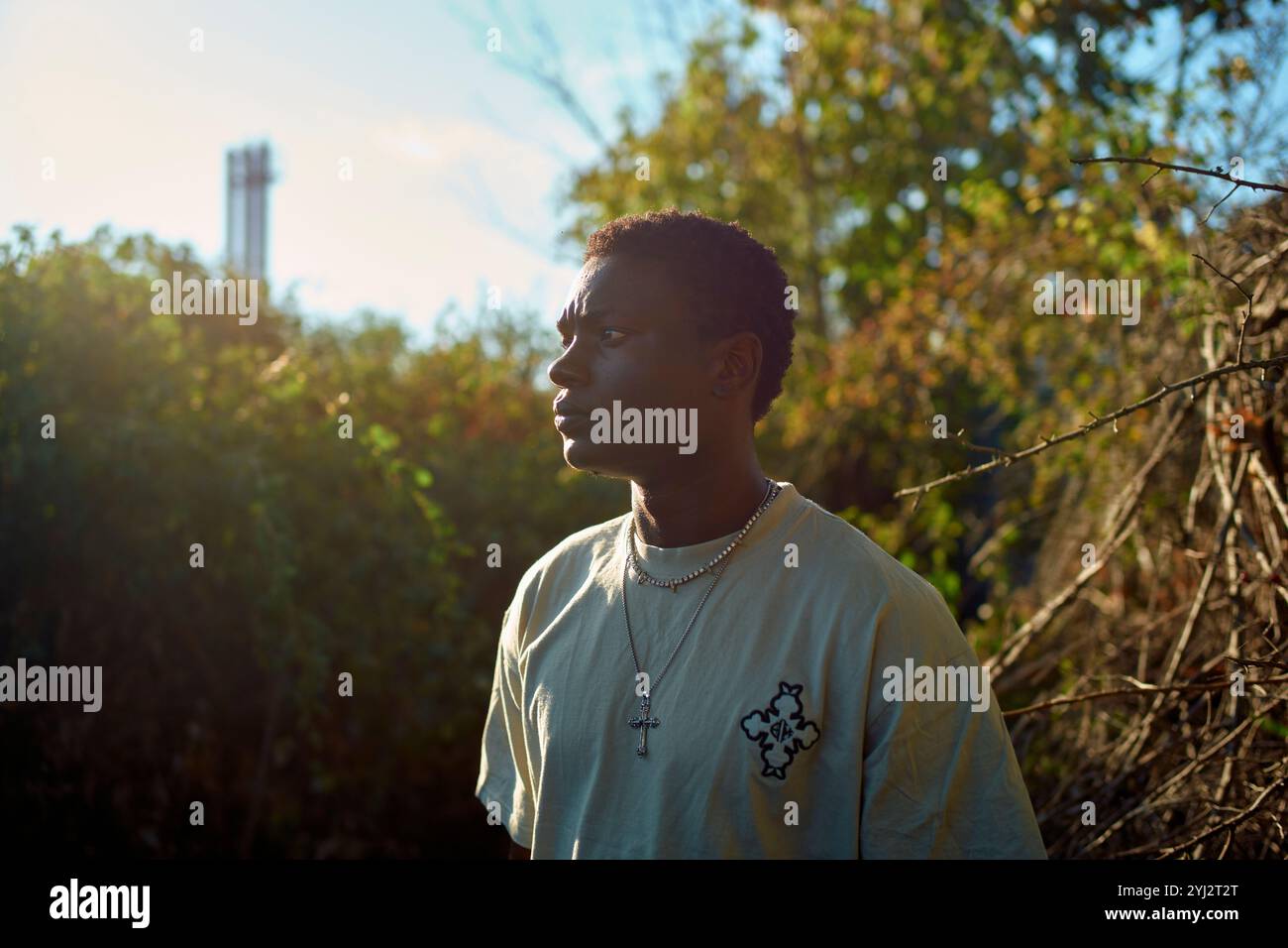 Un jeune homme pensif portant un collier se tient au milieu du feuillage avec la lumière du soleil projetant une lueur sur son profil, Berlin, Allemagne Banque D'Images