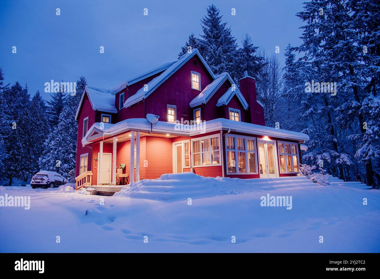 Maison rouge enneigée de deux étages avec fenêtres éclairées au crépuscule, entourée d'un paysage hivernal Banque D'Images