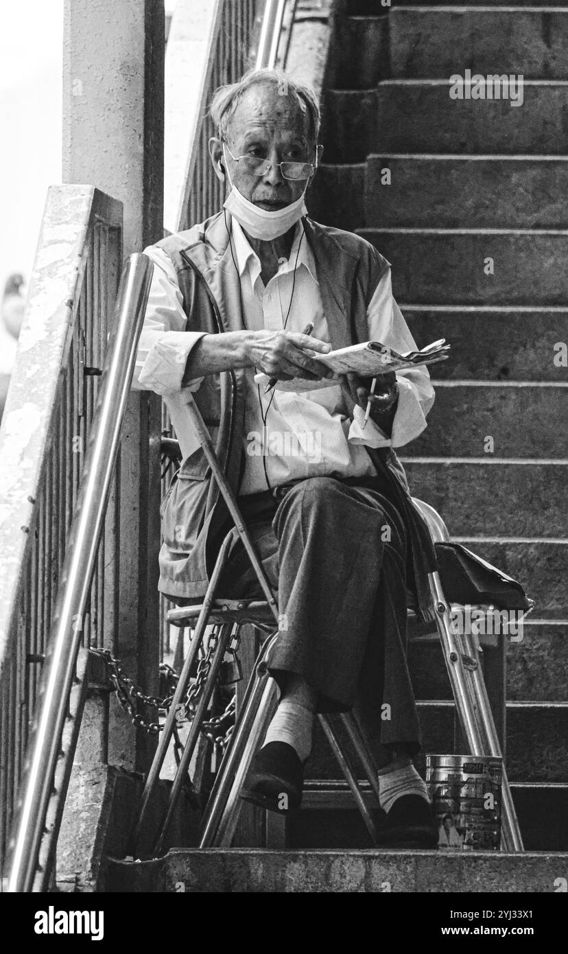 Un homme âgé est assis sur un escalier à Hong Kong, plongé dans la lecture d'un journal, mettant en scène un moment de calme au milieu de l'agitation urbaine. Banque D'Images