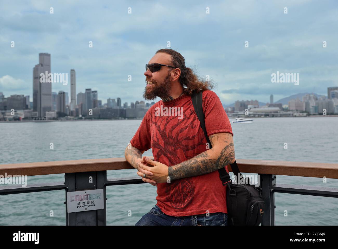 Un touriste se tient près de la rampe au bord de l'eau de Hong Kong, admirant l'horizon et le port par une journée claire et claire. Banque D'Images