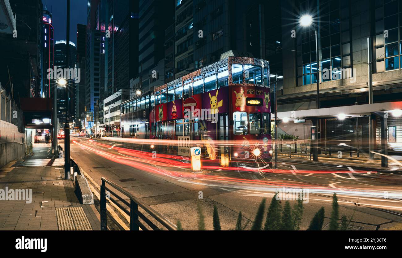 Un tramway animé se déplace le long d'une rue animée de Hong Kong, illuminé par les lumières de la ville et entouré de bâtiments modernes après la tombée de la nuit. Banque D'Images