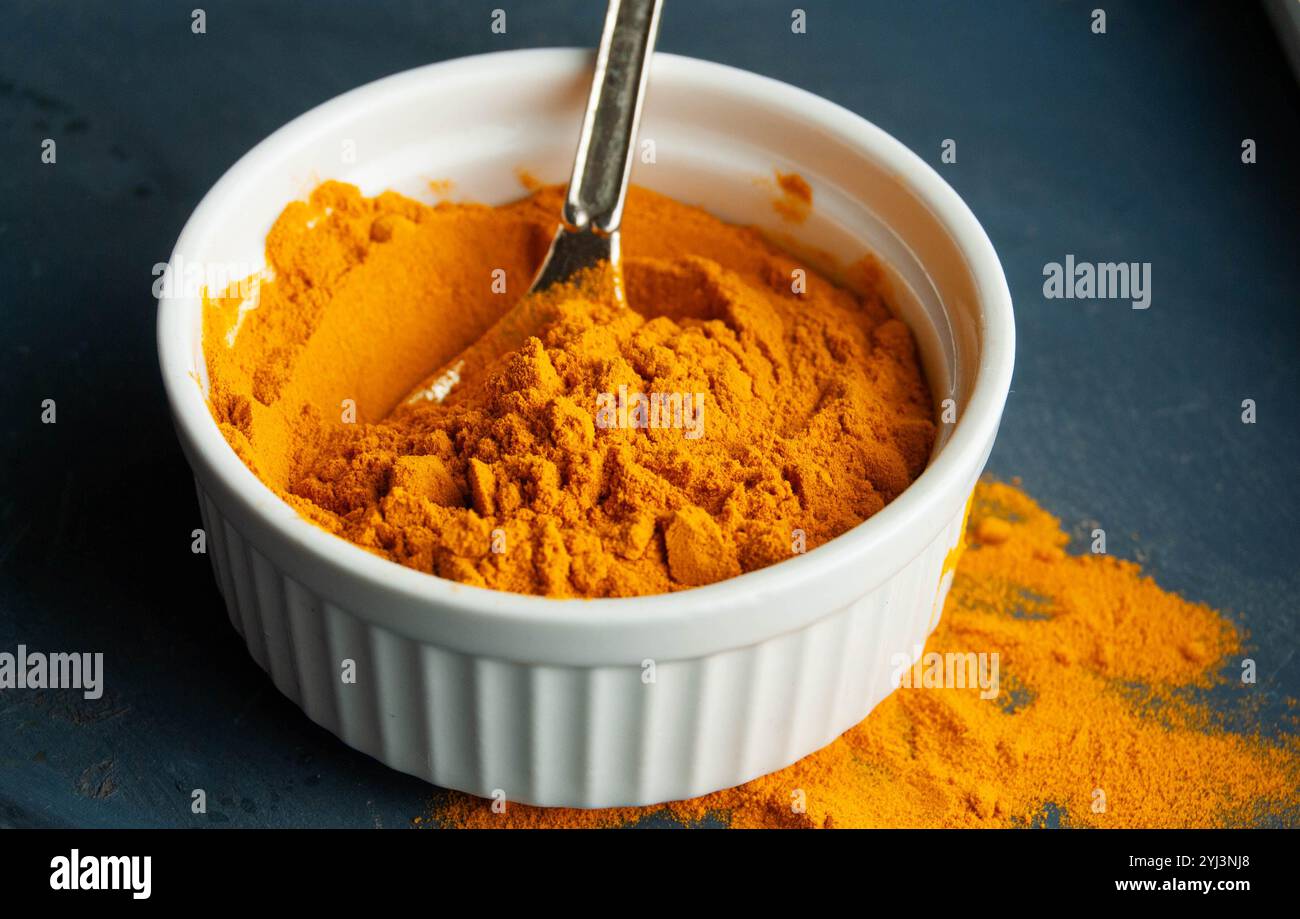 Poudre de curcuma brillante dans un petit bol blanc avec une cuillère sur fond bleu, soulignant des couleurs vibrantes. Banque D'Images