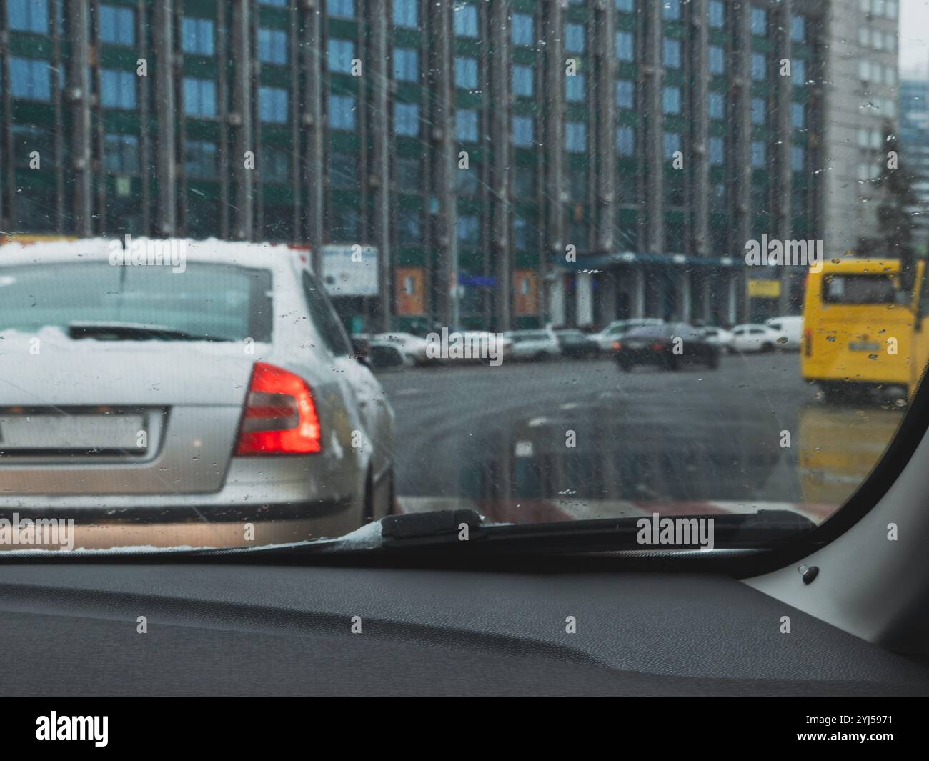 Vue depuis la cabine du conducteur sur la route avec des voitures et un bus jaune traversant le passage piéton par un jour gris pluvieux et enneigé. Bâtiment humide et route Banque D'Images