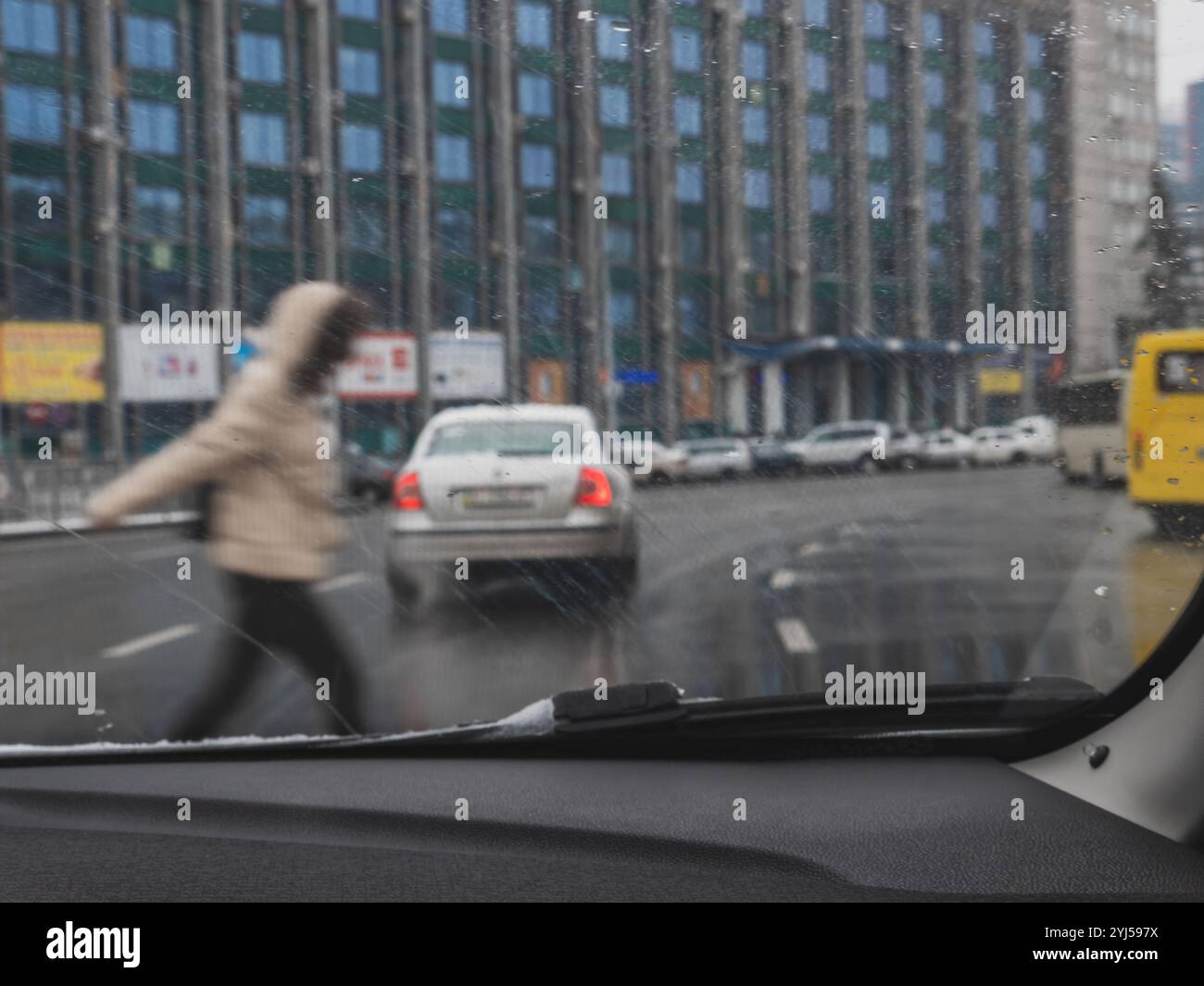 Vue depuis la cabine du conducteur sur les piétons traversant la route et les voitures et bus se déplaçant par un jour gris pluvieux et neigeux. Bâtiment et panneaux d'affichage à l'arrière Banque D'Images
