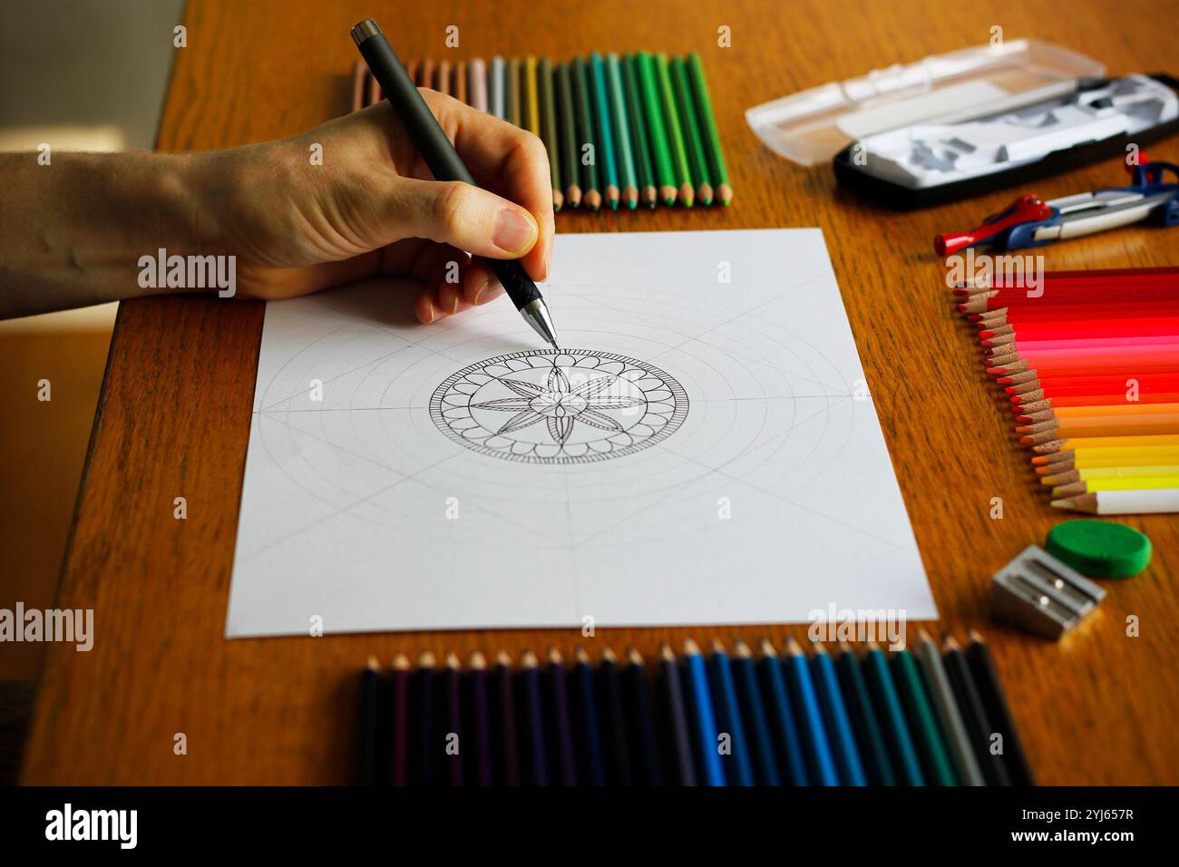 Main d'une femme caucasienne dessinant un motif mandala sur un papier blanc couché sur une table en bois, des crayons colorés et d'autres ustensiles de dessin à proximité Banque D'Images