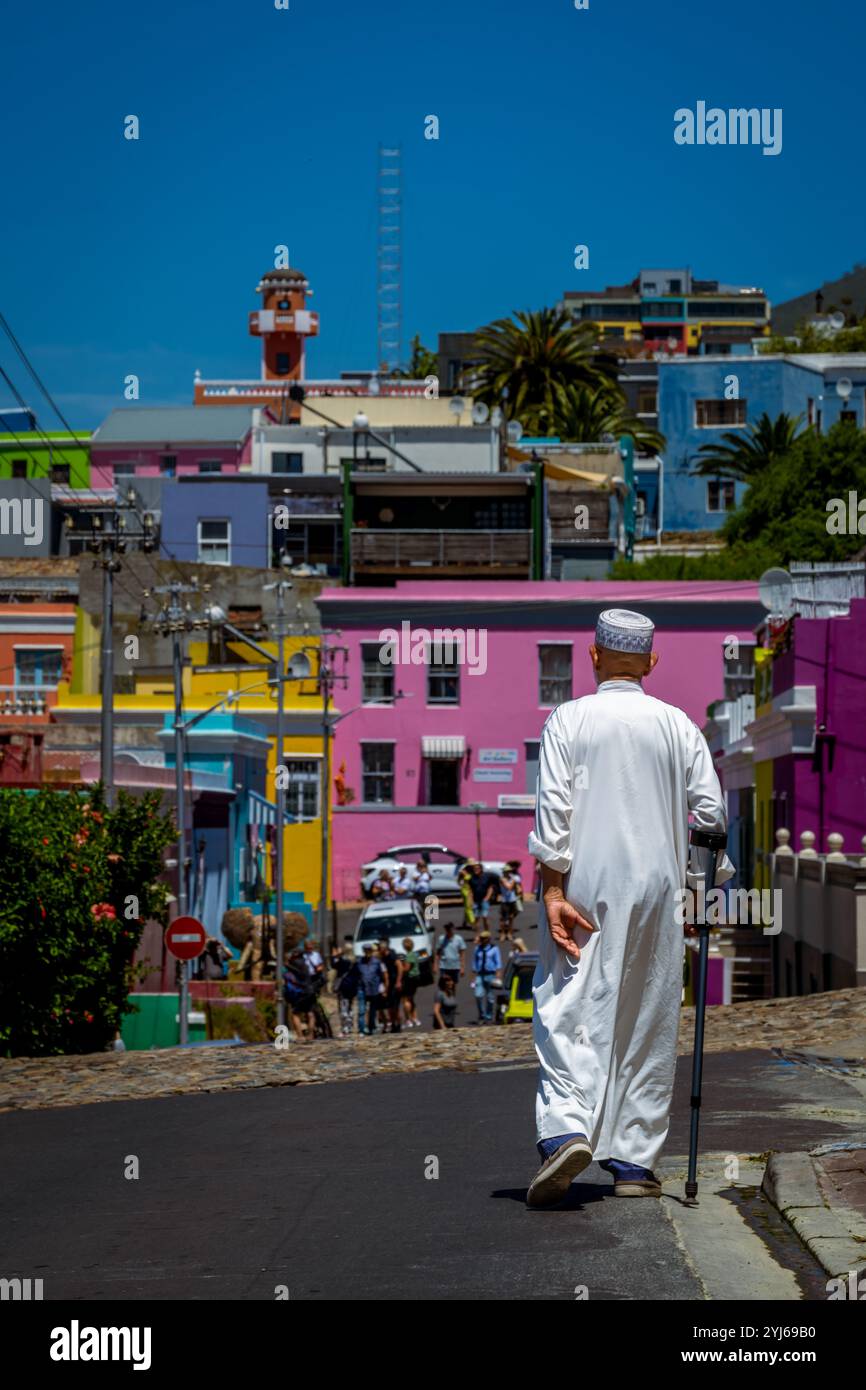 Un vieil homme musulman local dans une robe traditionnelle marchant avec une canne et des maisons colorées dans le quartier de Bo-Kaap au Cap, Afrique du Sud. Banque D'Images