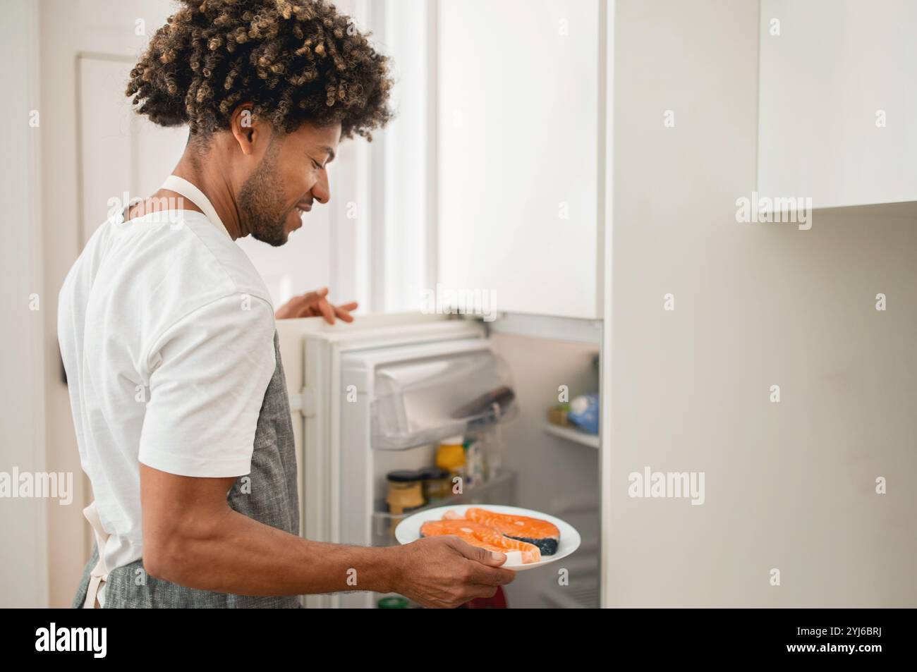 Un homme noir aux cheveux bouclés tient une assiette avec de la nourriture tout en atteignant son réfrigérateur. Il semble concentré sur la sélection des ingrédients pour son pré-repas Banque D'Images