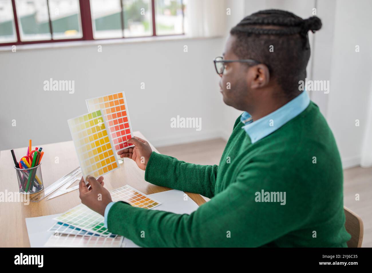 Un homme noir est assis à un bureau dans un espace de travail lumineux, comparant soigneusement les échantillons de couleur. Des crayons et des outils sont organisés à proximité, montrant son accent sur un d. Banque D'Images