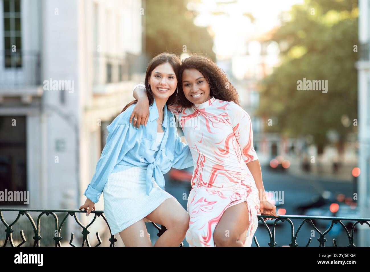 Deux jeunes femmes sourient et posent ensemble sur une balustrade dans un environnement urbain animé, profitant de la chaleur du soleil et de leur temps en plein air. Banque D'Images