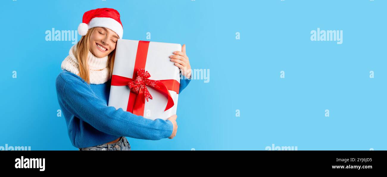 Femme caucasienne excitée dans le chapeau de Santa tenant et embrassant la grande boîte cadeau emballée sur fond bleu de studio. Heureuse dame célébrant le nouvel an ou Christma Banque D'Images