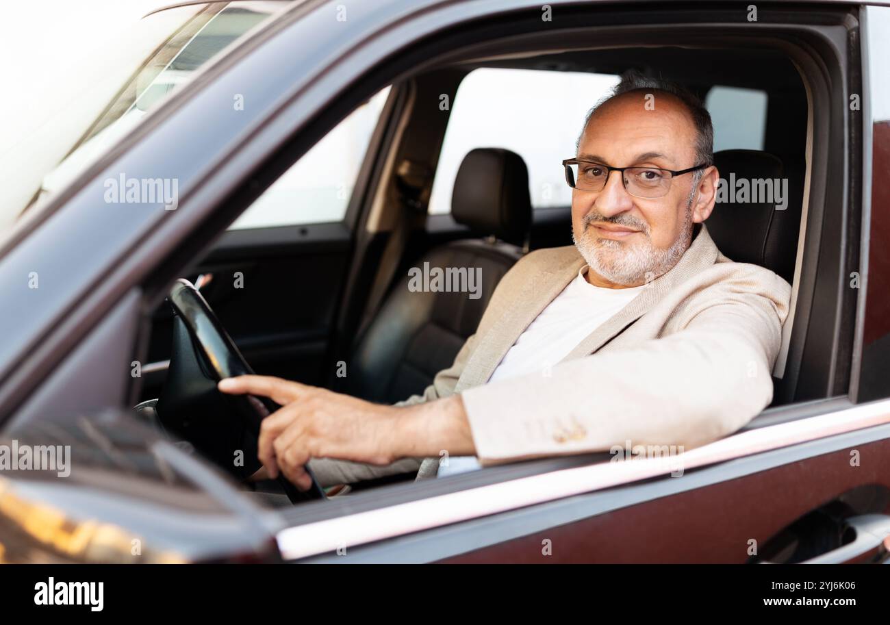 Un homme confiant avec des lunettes est assis dans le siège du conducteur d'une voiture, pointant vers l'extérieur pendant qu'il regarde la caméra. La lumière chaude du jour met en valeur son rel Banque D'Images