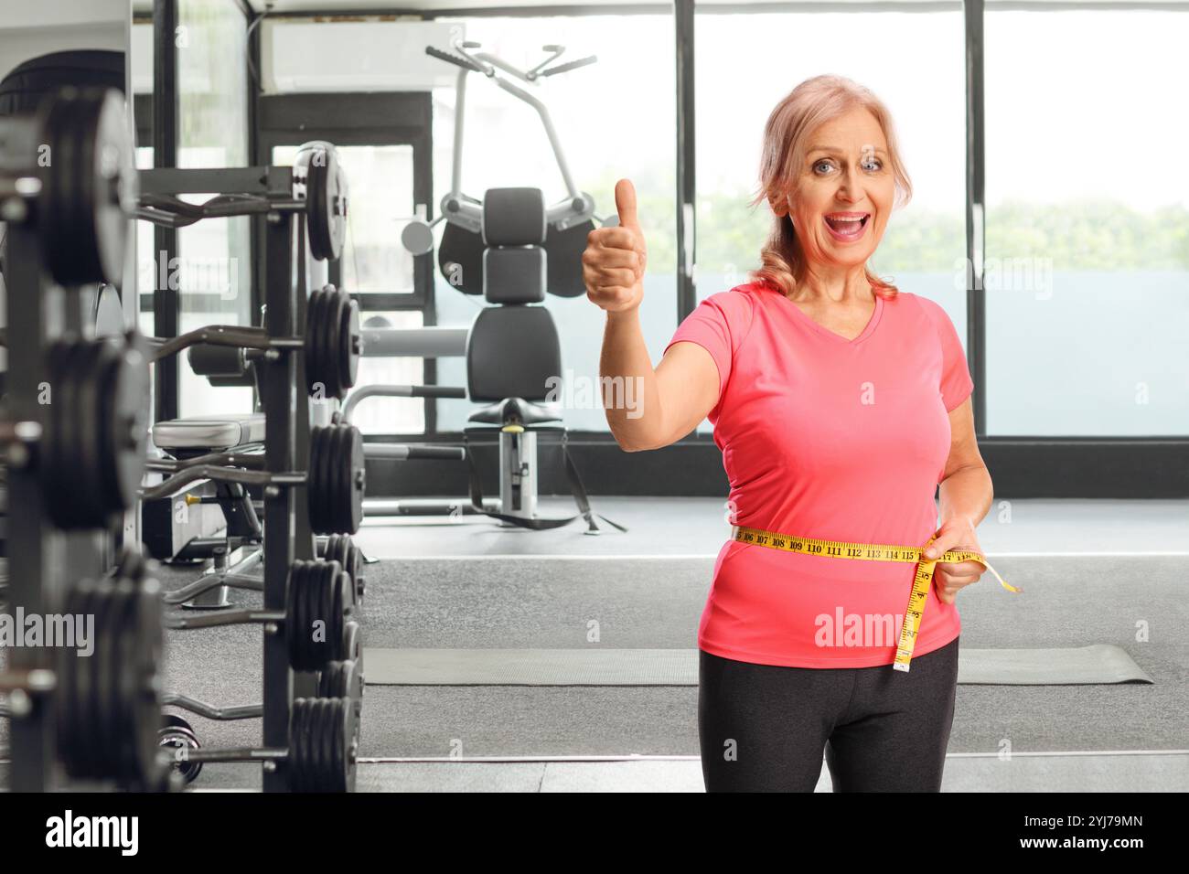 Femme mature mesurant sa taille avec une bande dans une salle de gym et faisant des gestes pouces vers le haut Banque D'Images