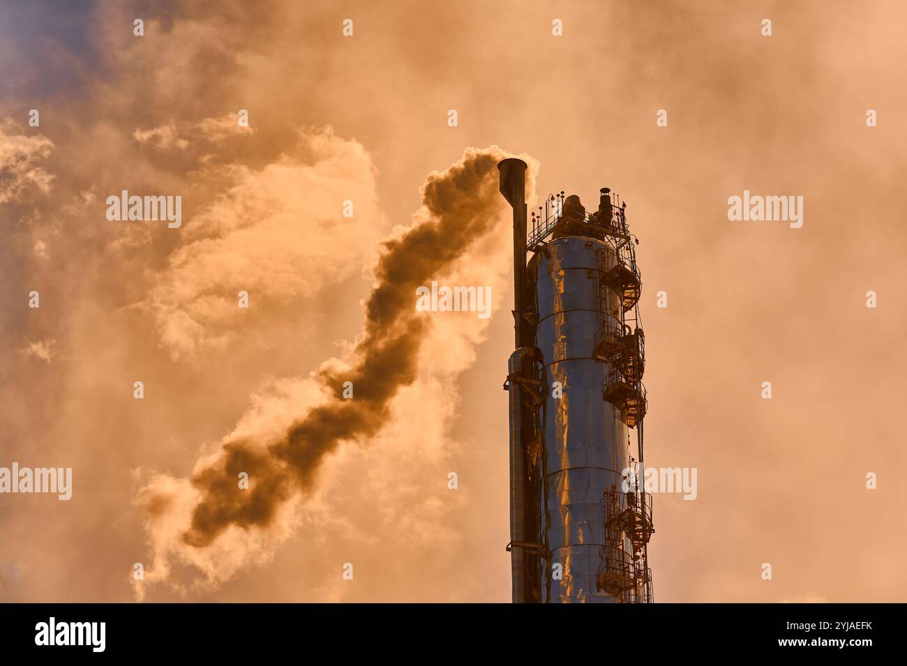 La colonne de distillation d'une usine chimique émet de la vapeur sous un ciel nuageux en fin d'après-midi dans un cadre industriel Banque D'Images