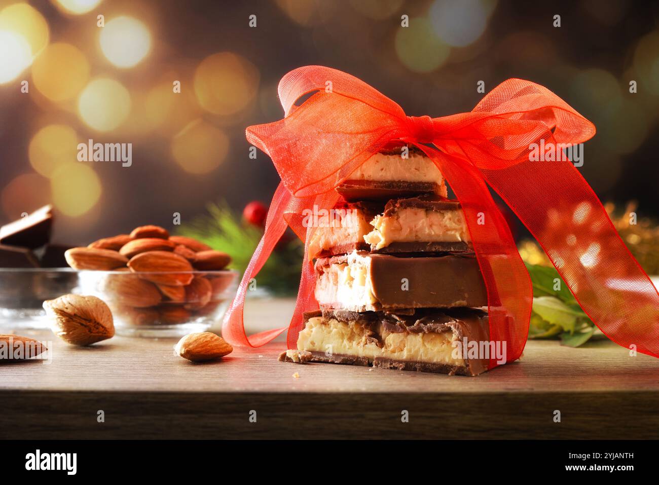 Nougat recouvert de chocolat avec des morceaux empilés attachés avec un ruban rouge sur une table en bois avec des amandes naturelles et des portions de chocolats et décors festifs Banque D'Images