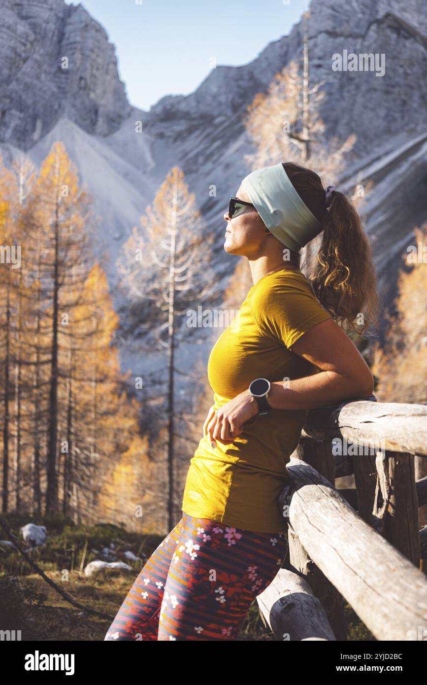 Vue latérale de jeune randonneur femme caucasienne dans des vêtements colorés, matériel de randonnée, appuyé sur une clôture en bois quelque part dans les montagnes, profitant de la vue Banque D'Images