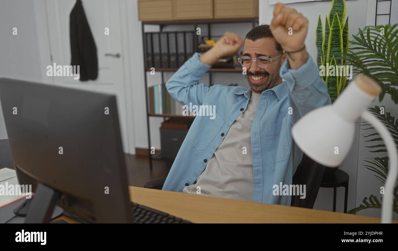 Jeune homme hispanique avec moustache célébrant dans un espace de travail de bureau avec une expression joyeuse. Banque D'Images
