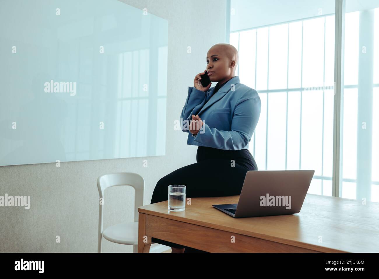 Une femme d'affaires afro-américaine confiante portant un élégant blazer bleu s'assoit sur un bureau dans un cadre de bureau contemporain. Elle passe un appel téléphonique Banque D'Images