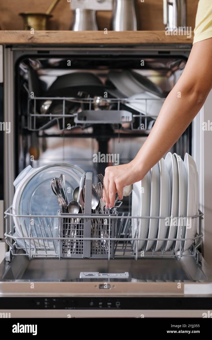 Vue avant de la main d'une femme enlevant les couverts propres d'un lave-vaisselle chargé. Fond de cuisine moderne Banque D'Images