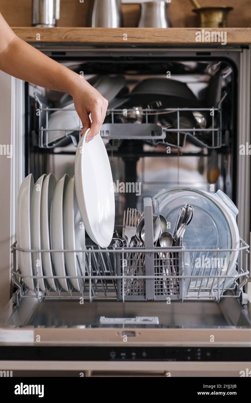Vue avant de la main d'une femme enlevant la plaque propre d'un lave-vaisselle chargé. Fond de cuisine moderne Banque D'Images