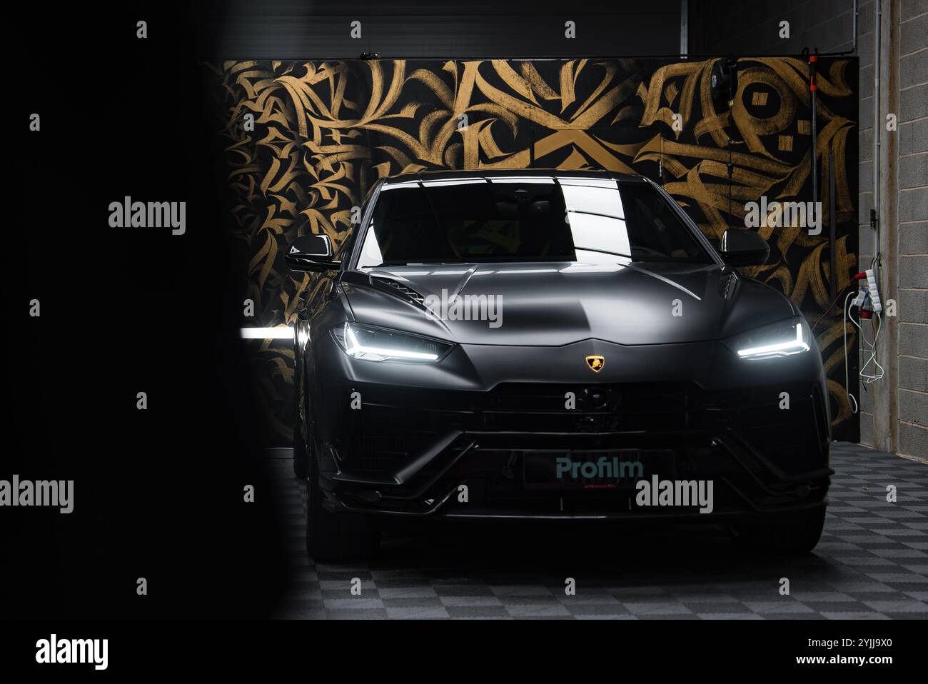 Une Lamborghini Urus performante noir mat est présentée à l’intérieur, mise en valeur par un éclairage ambiant. Des motifs dorés et un sol à carreaux rehaussent le luxu Banque D'Images