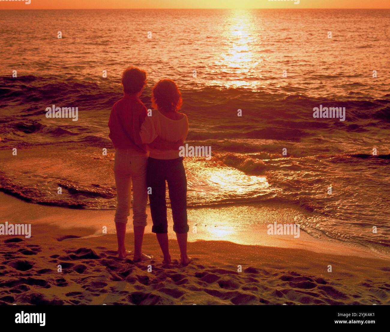 Vue arrière du jeune couple debout à la plage, regardant le coucher du soleil. Banque D'Images