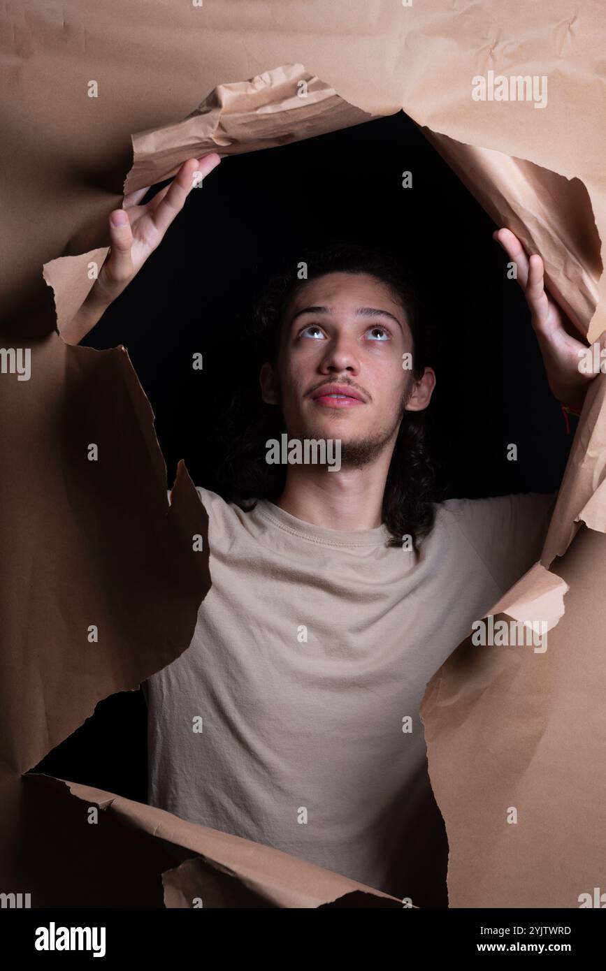 Un homme regardant à travers un trou dans une feuille de papier brun. Déchirant le papier avec ses mains. Portrait de studio. Banque D'Images