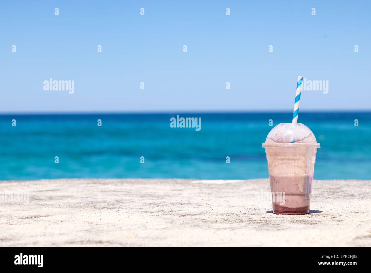 Milk-shake au chocolat à moitié plein avec paille bleue et blanche repose sur une surface sablonneuse au bord de la mer à Bronte Beach, Sydney, Australie. Banque D'Images