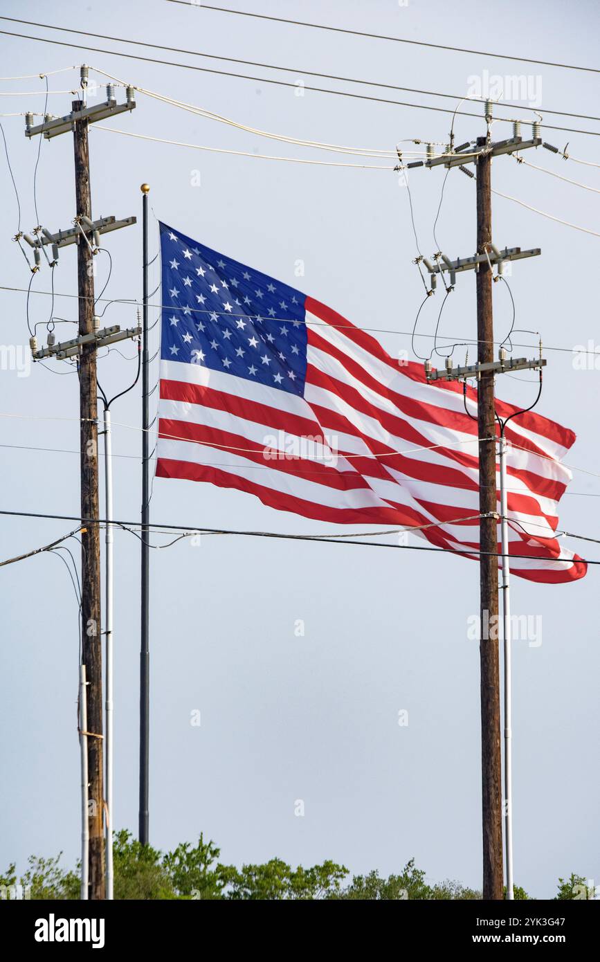 Drapeau AMÉRICAIN agitant au vent (à une distance de sécurité) derrière des poteaux électriques, dans le comté de Bexar, Texas, le 5 juin 2024. Fournir une électricité fiable et abordable est essentiel pour maintenir le bien-être économique et la qualité de vie de tous les résidents ruraux du pays. Le U/S. Département de l'Agriculture (USDA) développement rural (RD) rural Utilities Service (RUS) Programme électrique fournit leadership et capital pour maintenir, développer, améliorer et moderniser la vaste infrastructure électrique rurale de l'Amérique. En vertu de la loi sur l'électrification rurale de 1936, le programme électrique fait di Banque D'Images