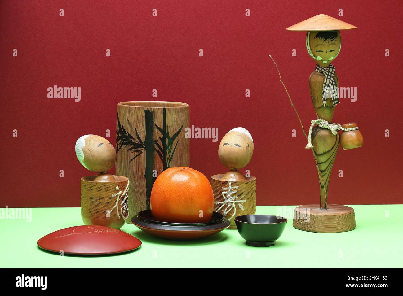 Photographie de natures mortes avec souvenirs – femme japonaise, deux poupées kokeshi, bambou peint, kaki sur fond de papier rouge. Gros plan avec Banque D'Images