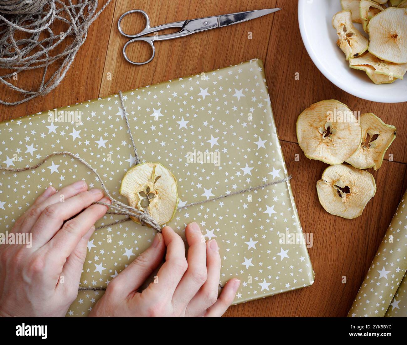 Mains féminines enveloppant le cadeau de Noël. Cadeau de Noël enveloppé dans du papier doré et décoré avec pomme séchée, pommes séchées dans un bol, ruban et ciseaux Banque D'Images