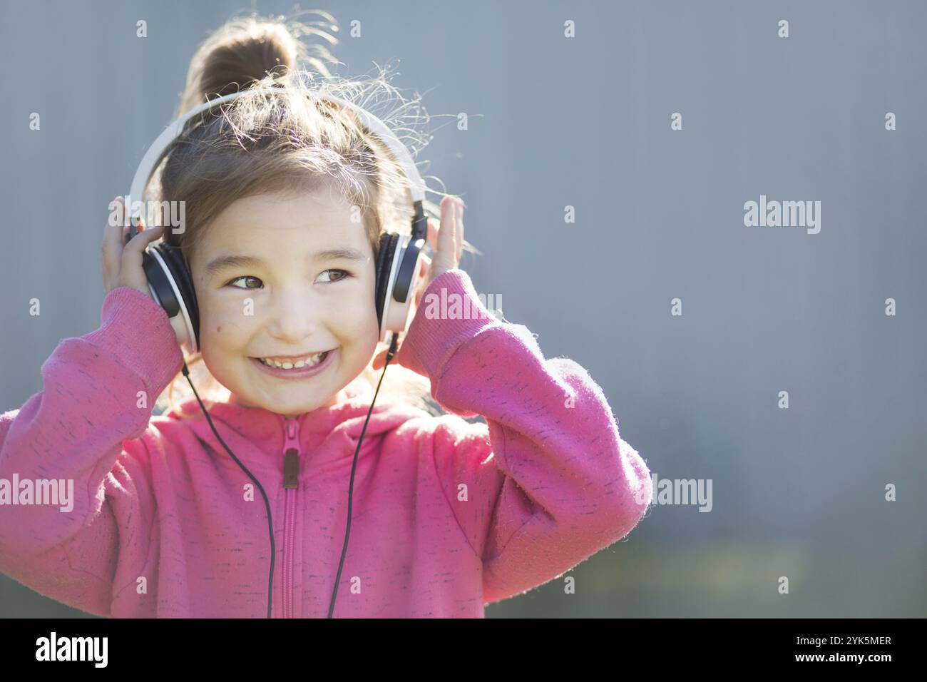 Une jeune fille gaie dans un casque et un pull à capuche rose écoute de la musique et rit. Extérieur, fond gris Banque D'Images