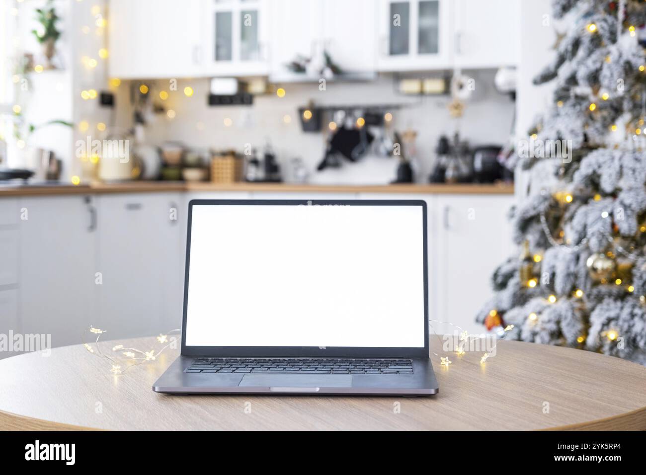 Ordinateur portable avec une maquette d'écran blanc dans la cuisine de Noël décorée blanche confortable avec des lumières de fée et un arbre de Noël. Télétravail saisonnier, interne Banque D'Images