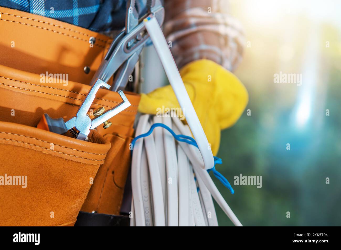 Un électricien se tient à l'extérieur, tenant des câbles blancs enroulés et une clé dans une ceinture à outils, prêt pour une tâche pendant la journée. Banque D'Images