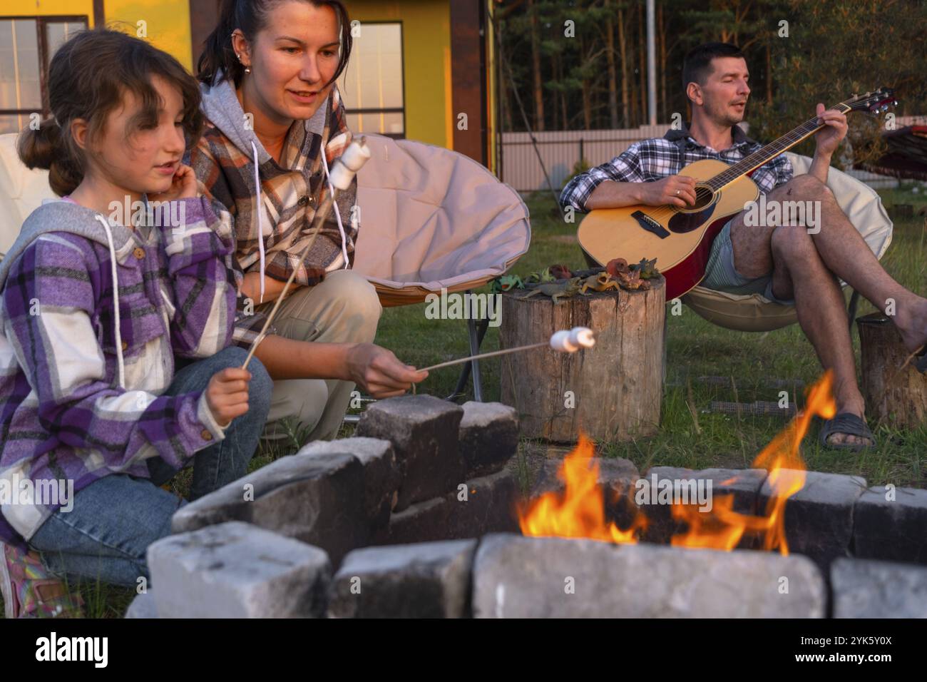 Une fille dans une chemise à carreaux rôtit des guimauves sur un feu dans la cour de la maison, père joue de la guitare. Soirée de réunion en famille près du feu de camp, Banque D'Images