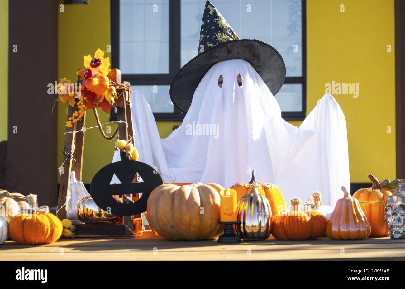 Enfant dans le drap de lit avec des fentes comme fantôme dans le chapeau de sorcière et le décor d'Halloween sur le porche de la maison à l'extérieur dans la cour de citrouille, lanterne, guirlande Banque D'Images